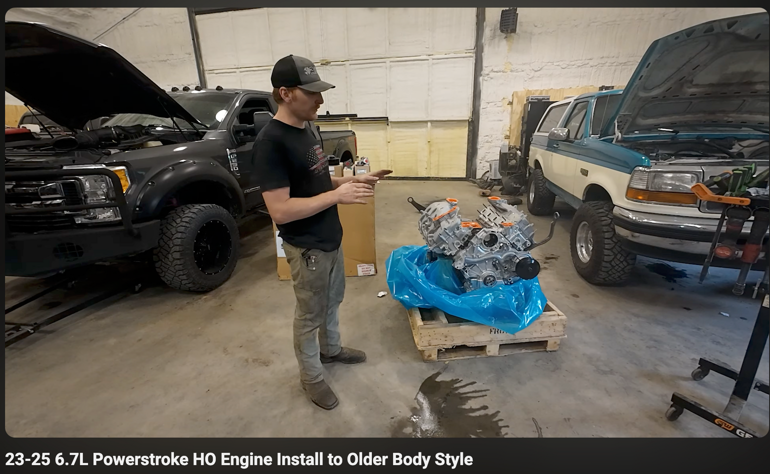 Man in a garage with automotive engine and vehicles with hoods up.