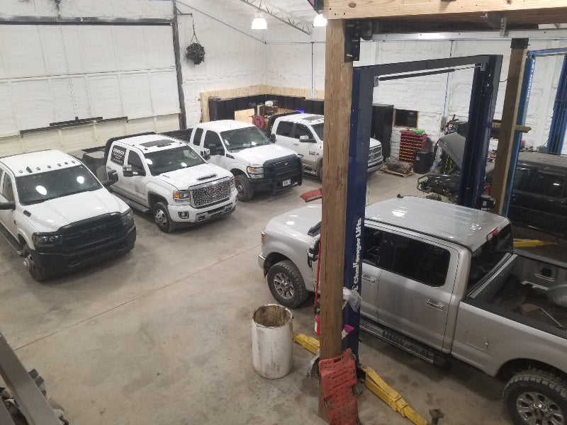 Garage with multiple trucks, including a white truck being lifted on a hydraulic lift.